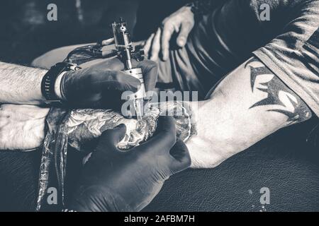 Tattoo artist in black gloves draws new black tattoo with tattoo machine on client`s male arm. Toned black and white. Stock Photo